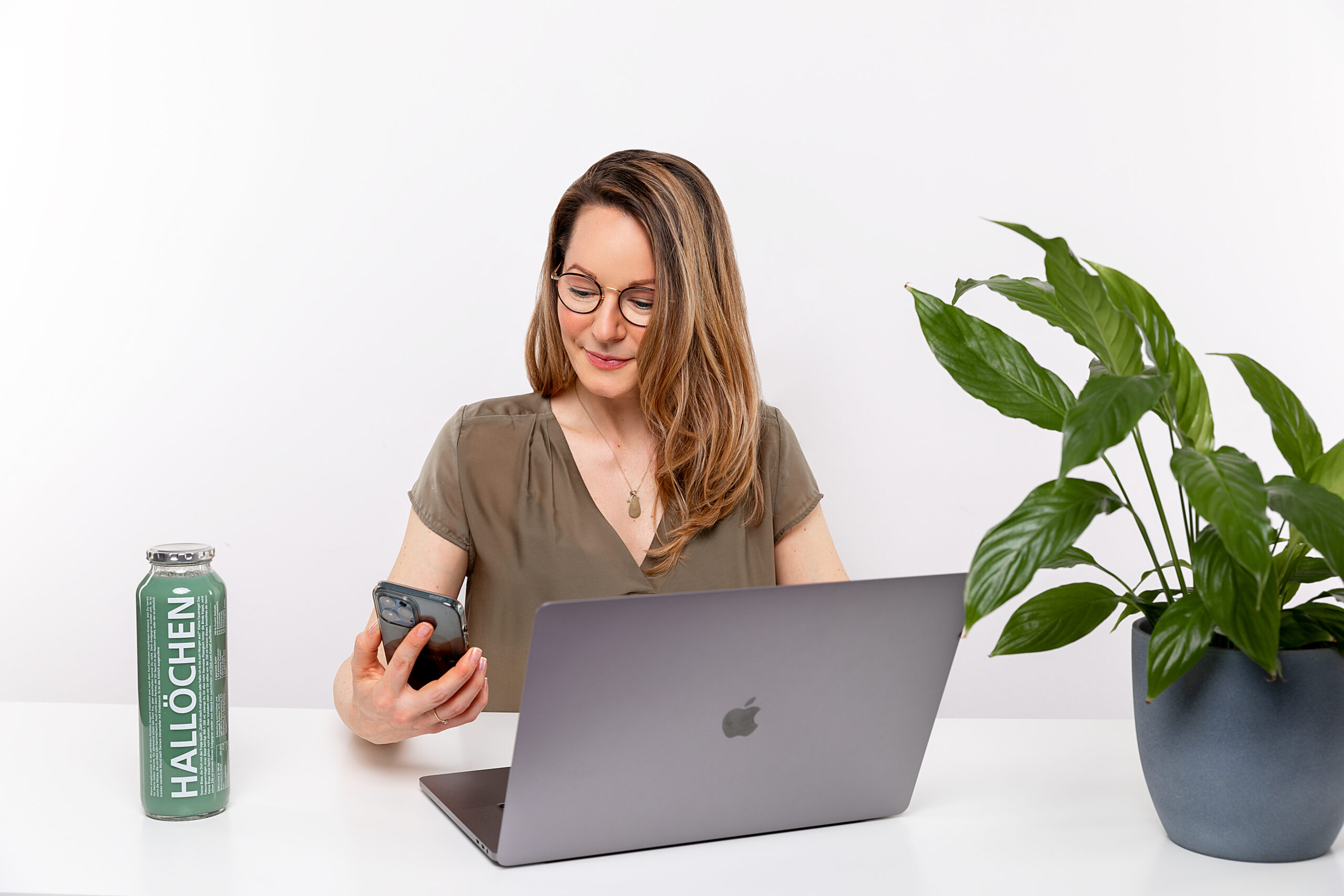Vorstellung von Christiane Dölz. Frau sitzt am Tisch mit Laptop vor ihr, einer Pflanze rechts von ihr. Links von ihr steht eine Glasflasche mit dem Aufdruck Hallöchen. Die Frau hält ein Handy in der Hand, auf das sie gerade schaut.